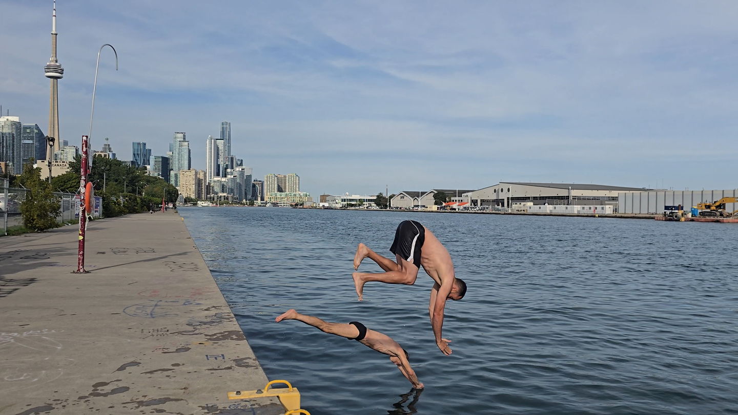 Swimmers entering the water at the west end of the pier, a yellow ladder handrail visible at the edge of the deck