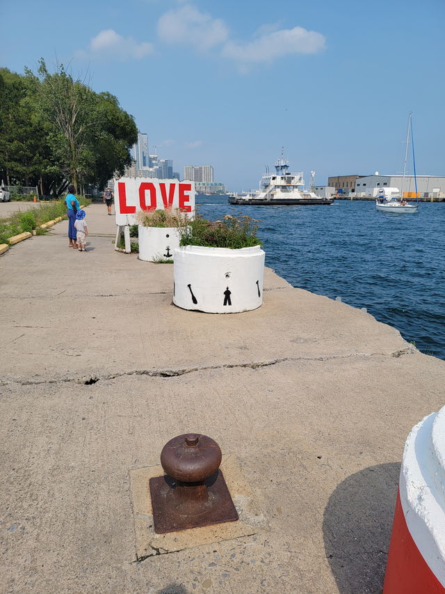 A rusted mooring bollard set into the pier deck, with the LOVE installation and a ferry in the distance